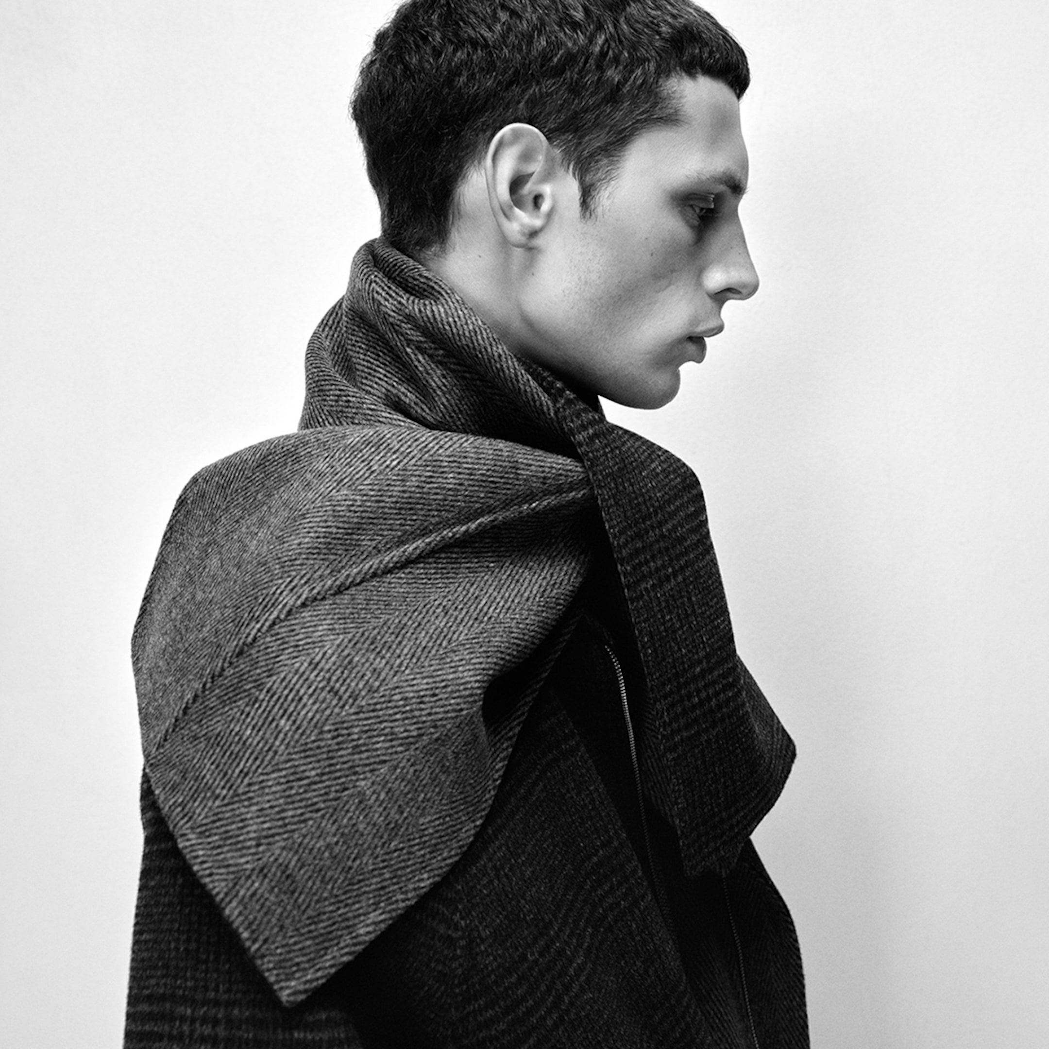 Side profile of a person with short hair, wearing a textured, plaid-patterned scarf wrapped around their neck, in black and white.