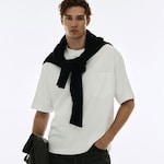 Placeholder for: Man in a white t-shirt with a black sweater draped over his shoulders, standing with one hand in pocket against a plain background.