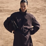Placeholder for: Person in a brown coat stands confidently with one hand on hip, against a sandy outdoor background.