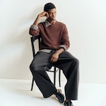Placeholder for: A person sits on a chair wearing a brown sweater over a gray shirt, with wide-legged black pants and black shoes, striking a thoughtful pose.