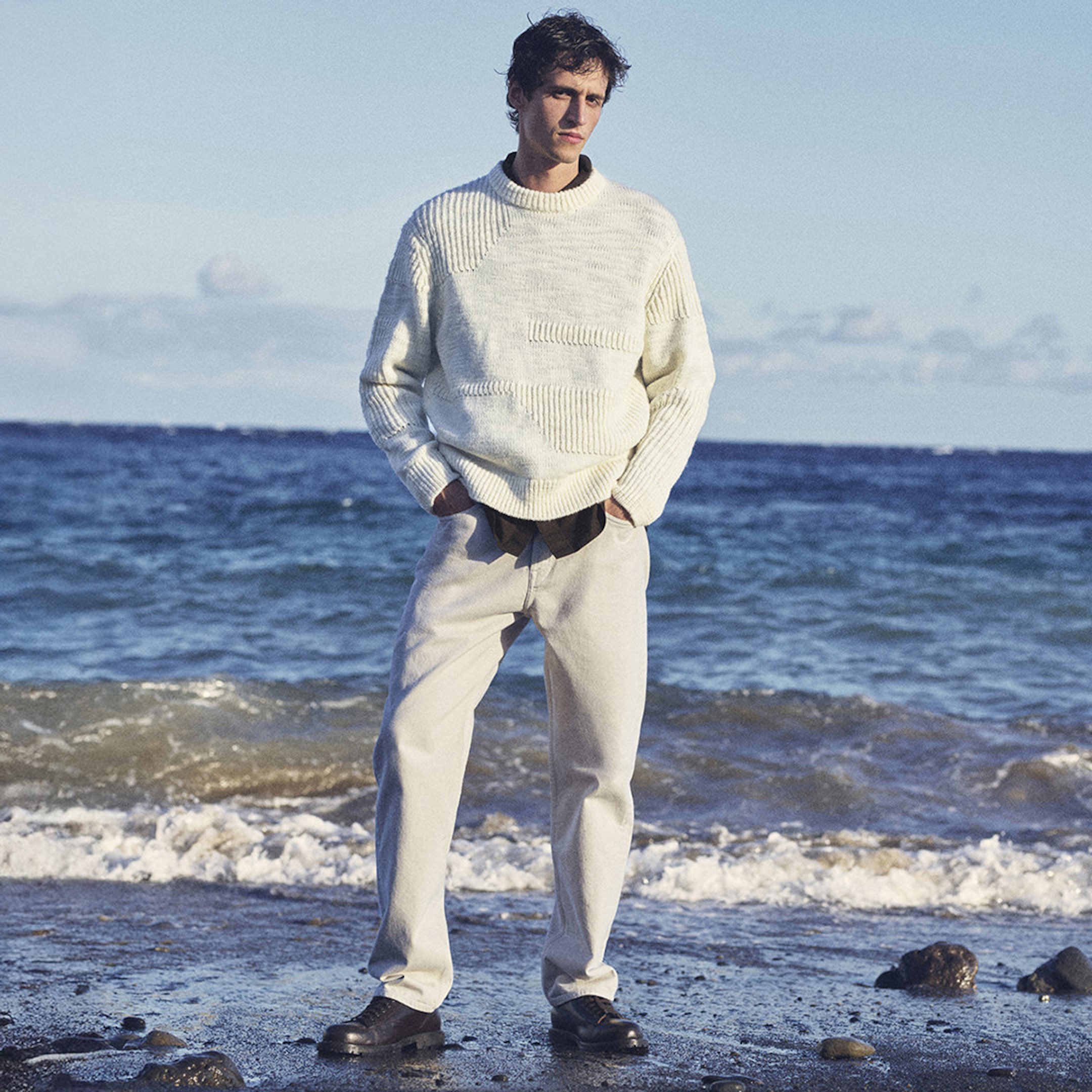Man in a white sweater and light pants standing on a beach with waves in the background under a clear sky.