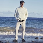Placeholder for: Man in a white sweater and light pants standing on a beach with waves in the background under a clear sky.