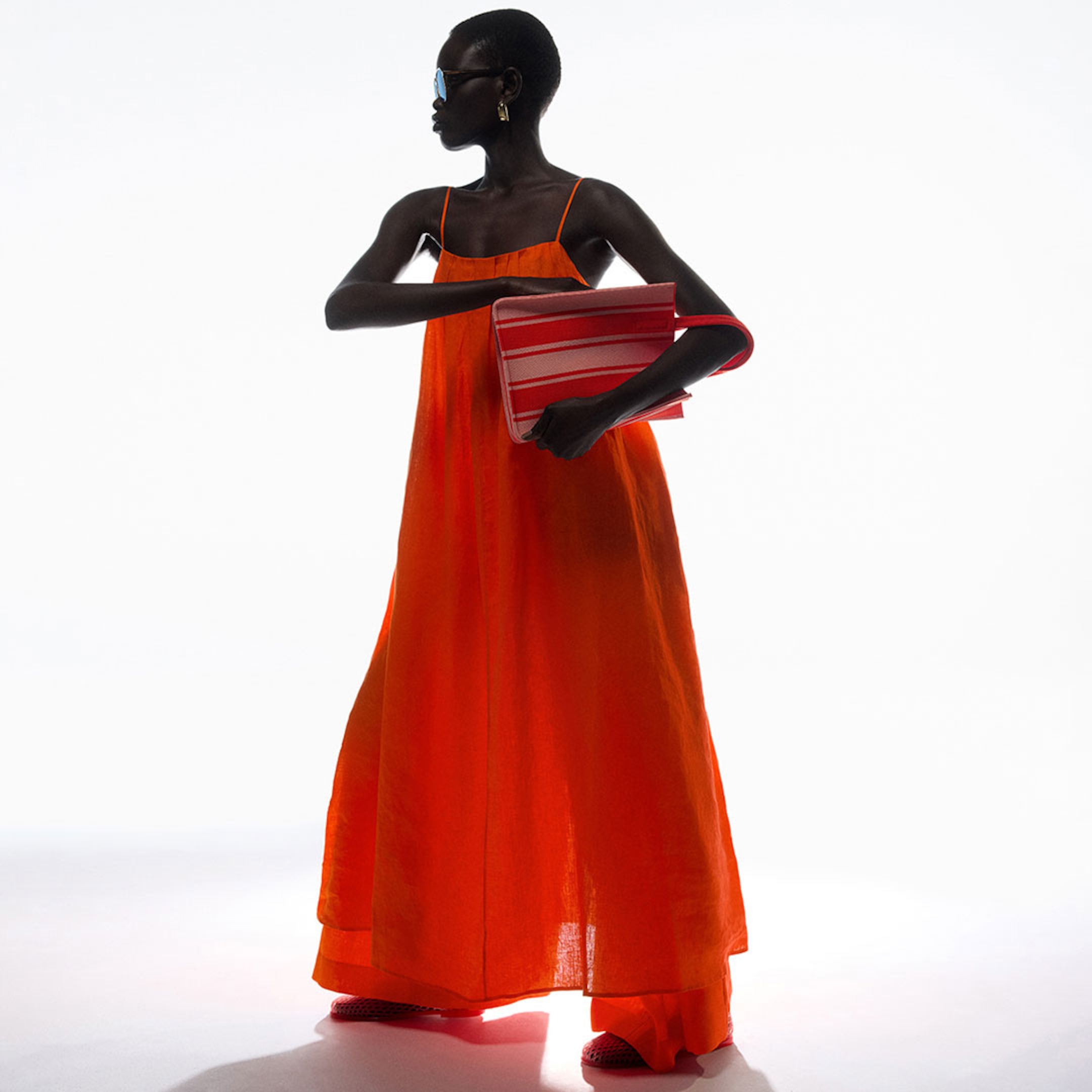Person in an orange dress and sunglasses holds a red-striped bag, standing against a bright white background.