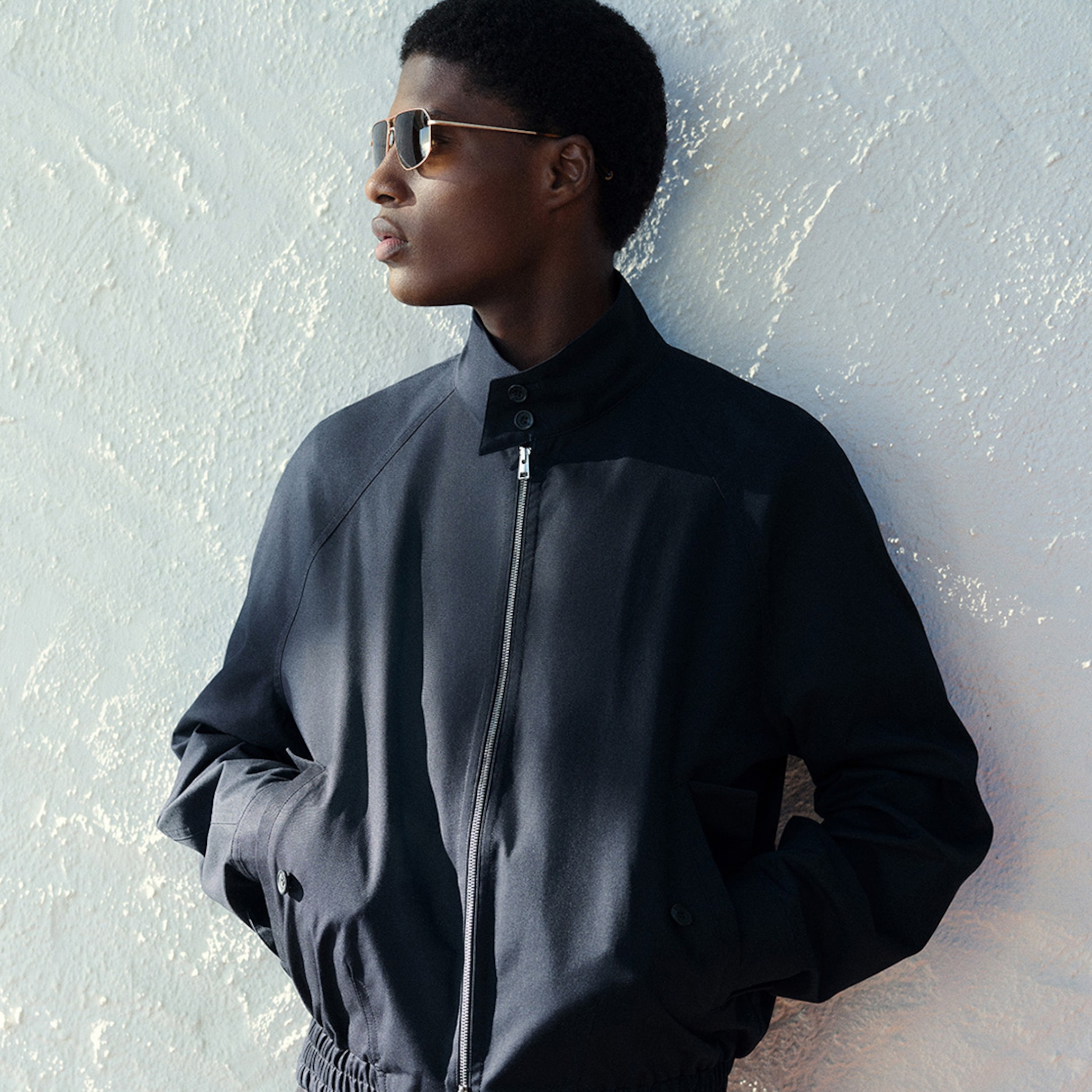 Person in a dark jacket and sunglasses leans against a textured wall, gazing to the side with hands in pockets.