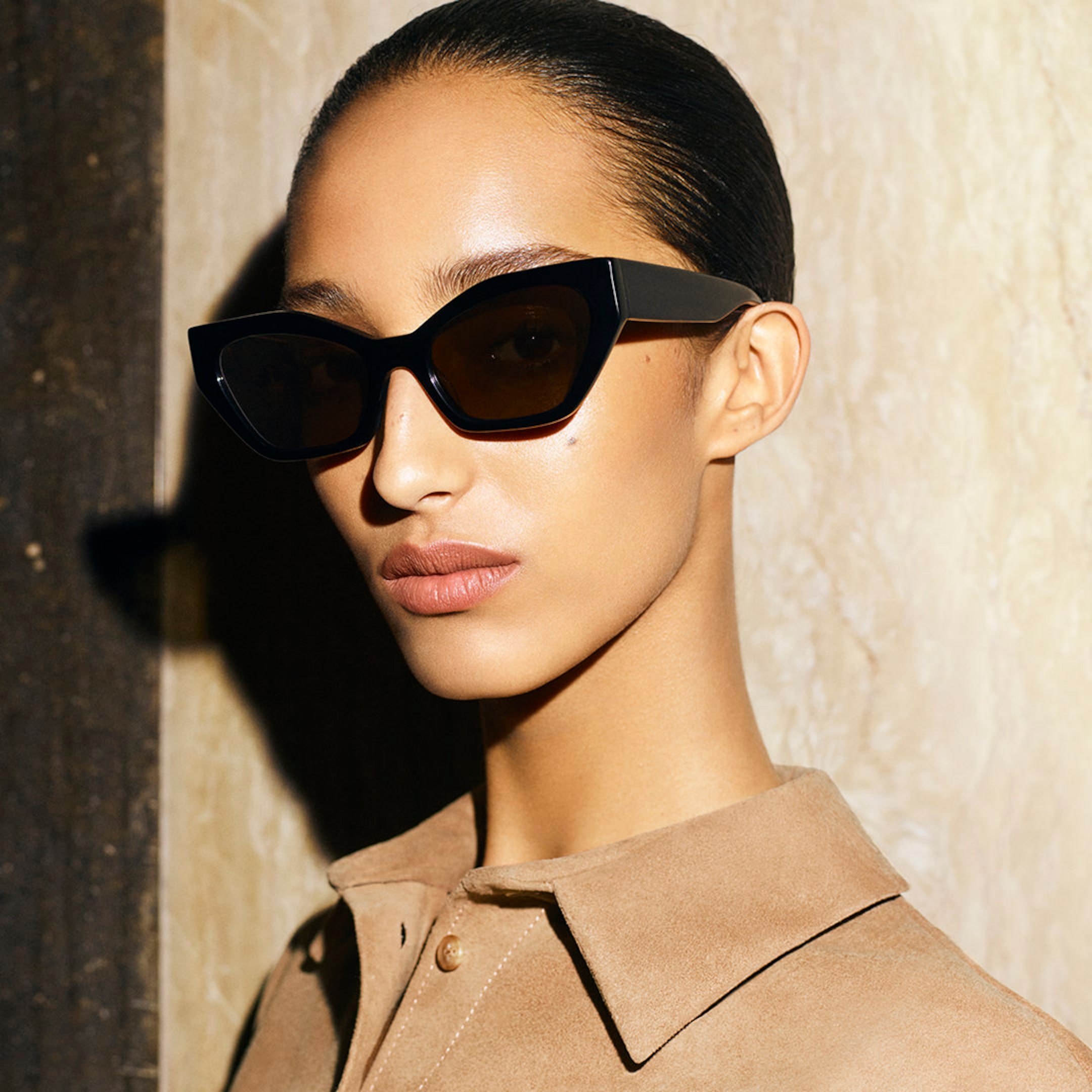 Woman with sleek hair wearing black cat-eye sunglasses and a beige collared shirt, standing against a textured wall.