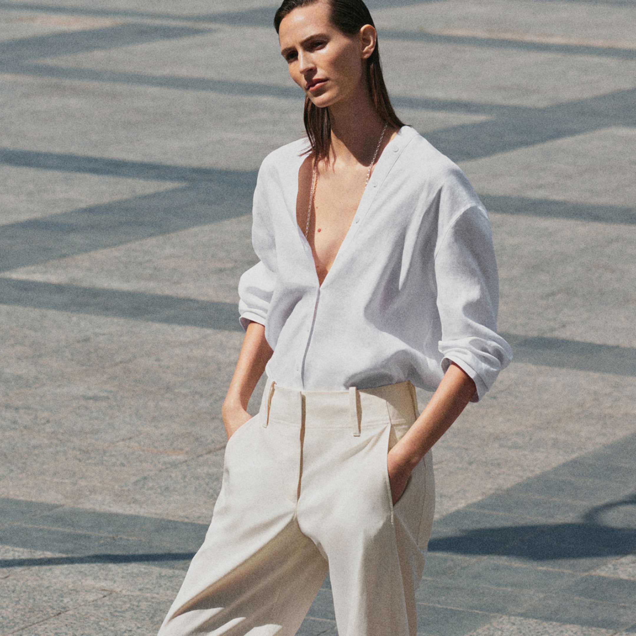 Person wearing a loose white shirt and cream trousers stands confidently in a sunny, open paved area.