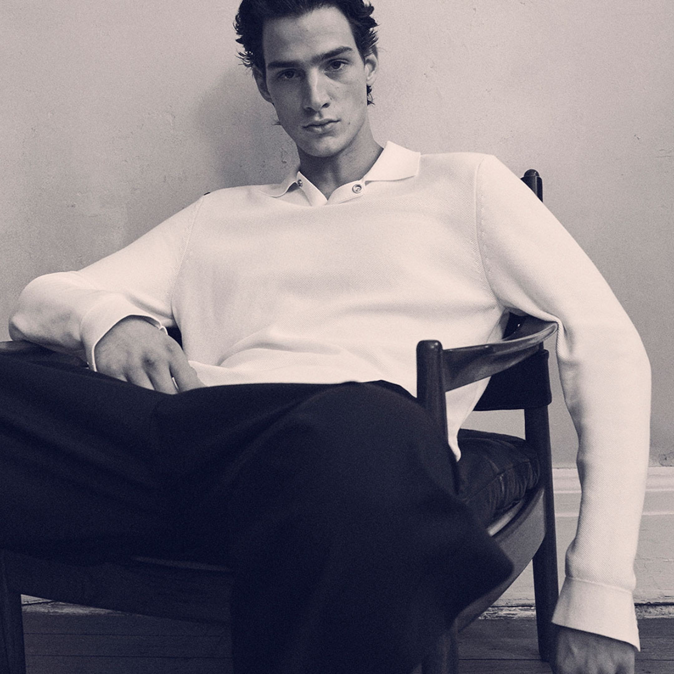 Man in a white polo shirt sits casually in a chair, leaning back with legs apart, against a plain wall.