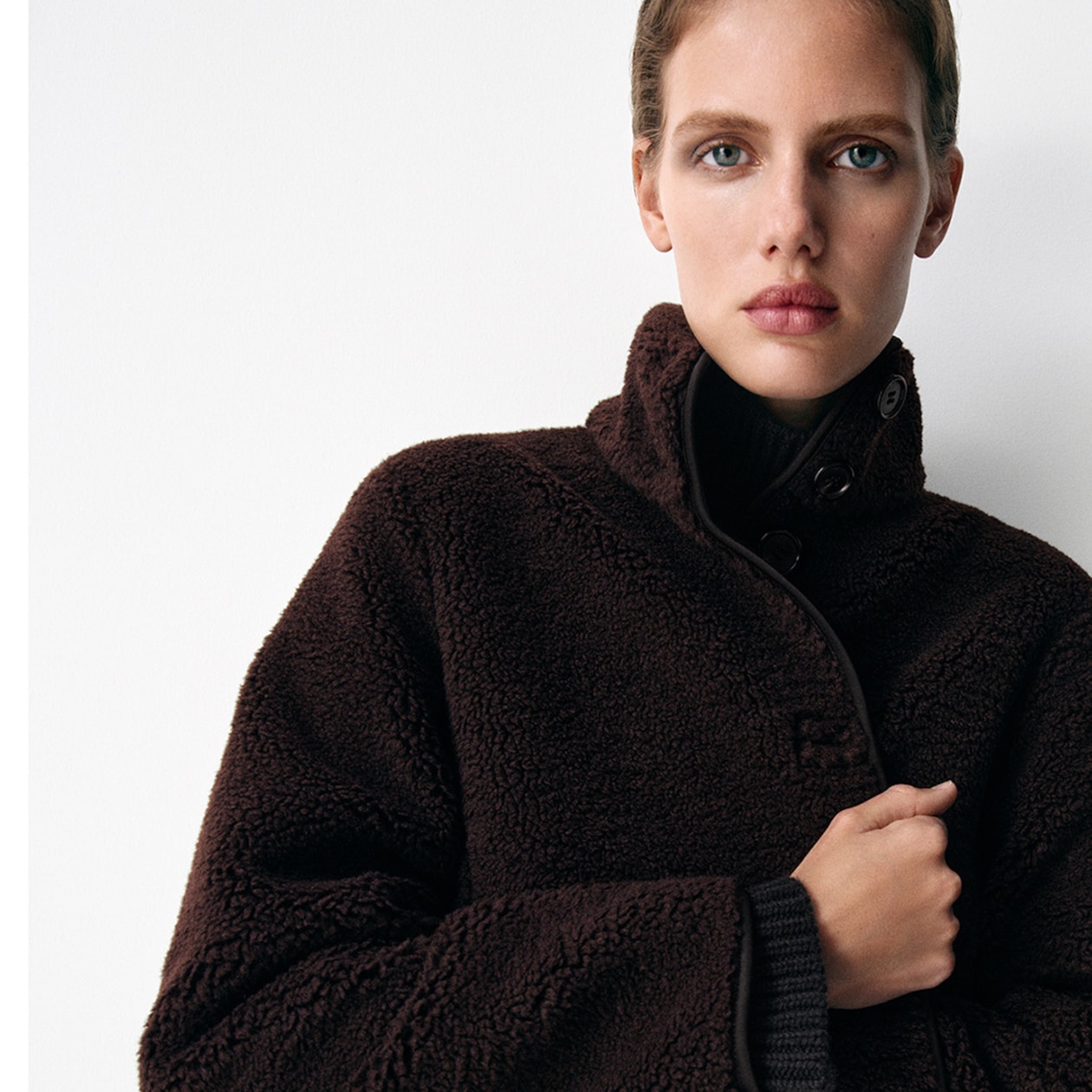 Person wearing a dark brown wool coat with button details, looking directly at the camera against a plain white background.