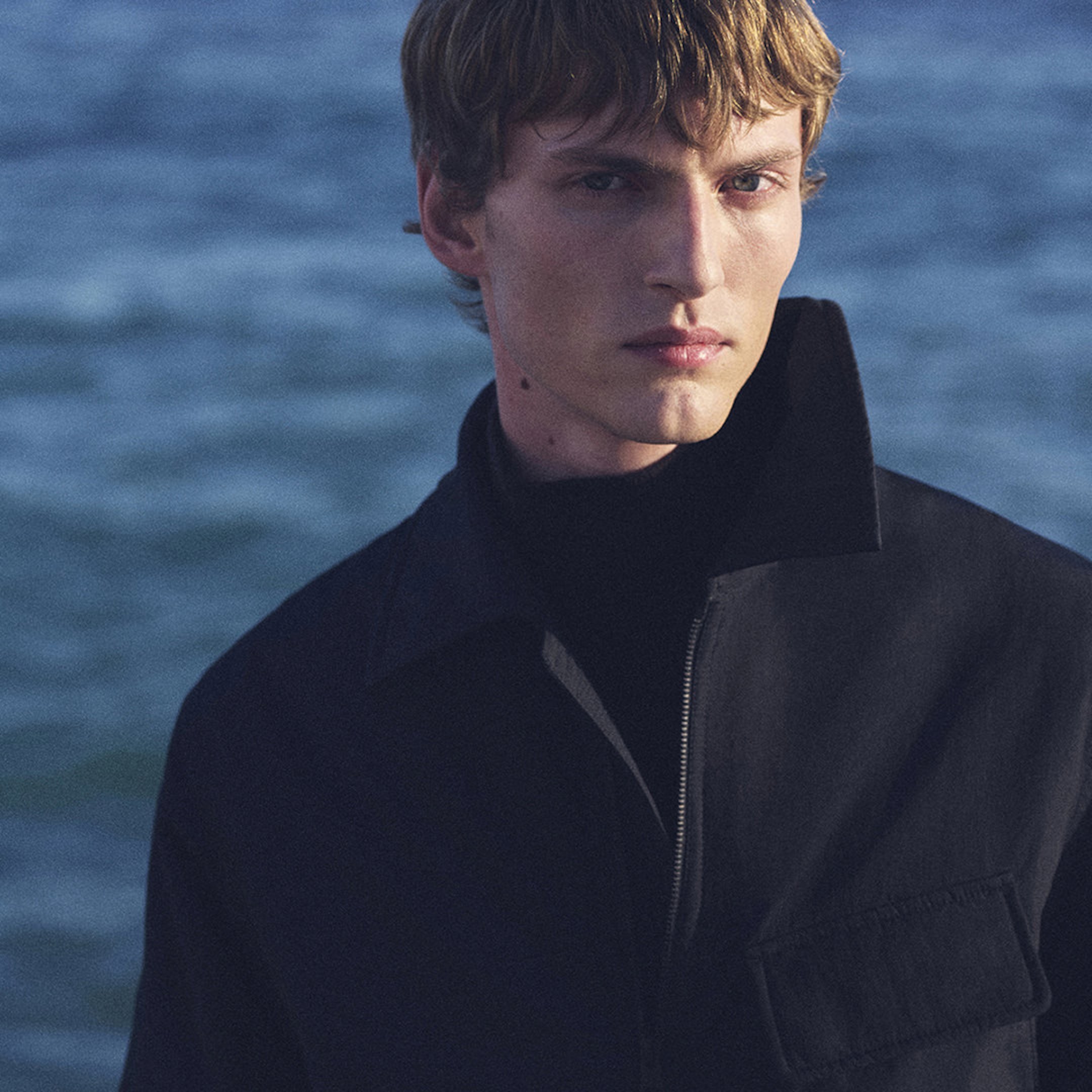 A person with short hair wearing a dark jacket, standing by a body of water with a calm expression.