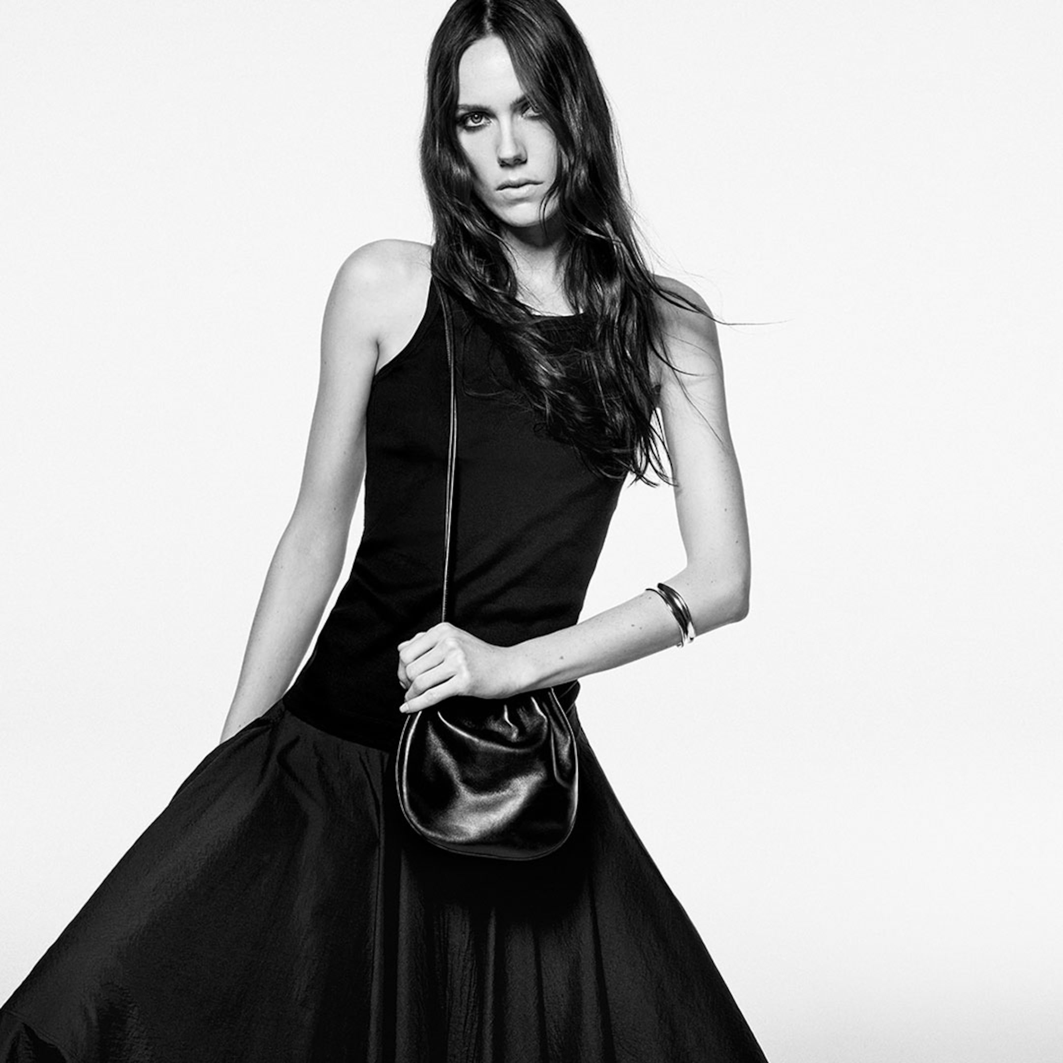 Woman in a black tank top and skirt poses with a crossbody bag against a plain background, exuding a confident and stylish vibe.