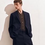 Placeholder for: Man in a dark suit and brown shirt leans against a wall, looking to his left.
