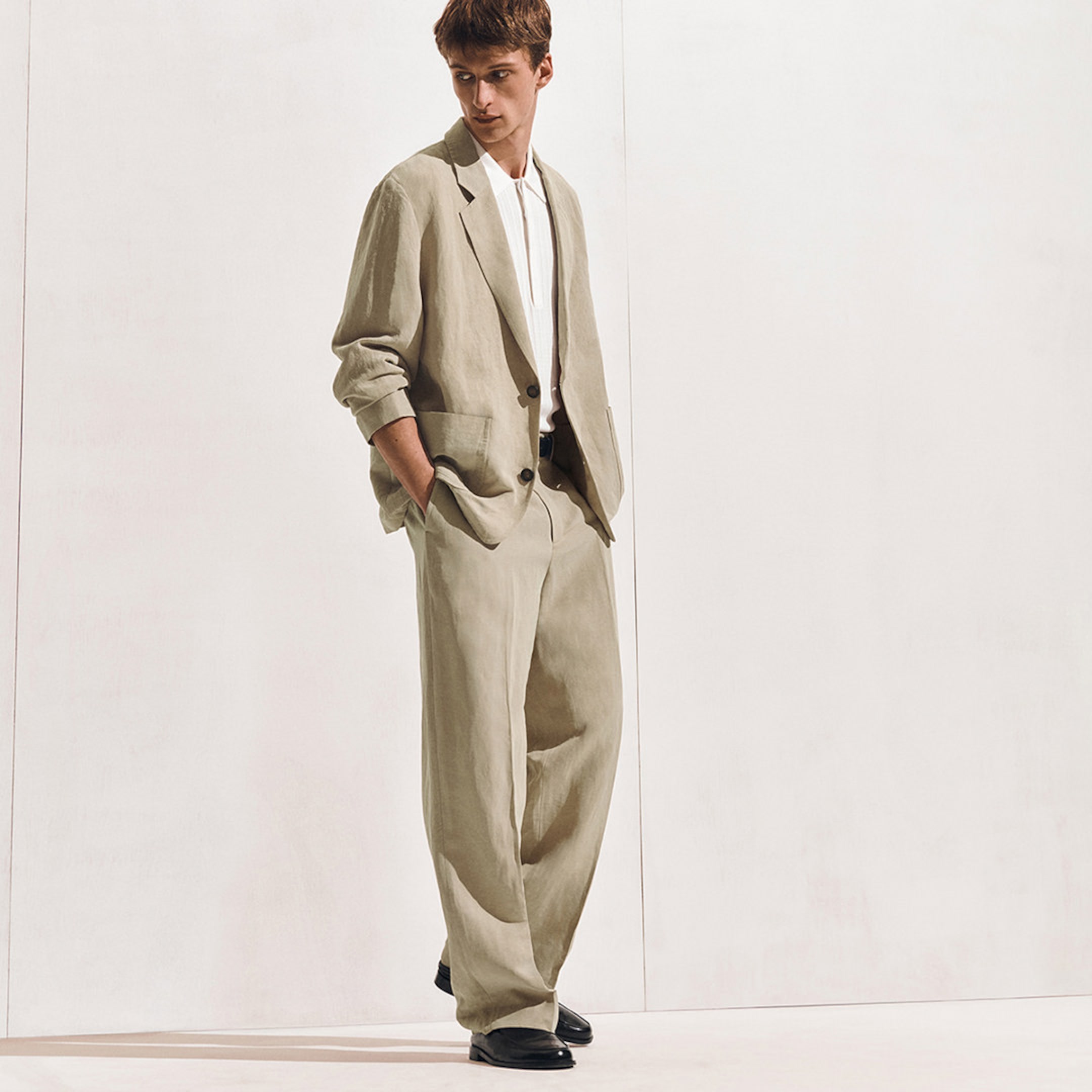Man in a stylish beige suit with white shirt, standing against a plain background, hands in pockets, wearing black shoes, looking down.