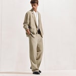 Placeholder for: Man in a stylish beige suit with white shirt, standing against a plain background, hands in pockets, wearing black shoes, looking down.