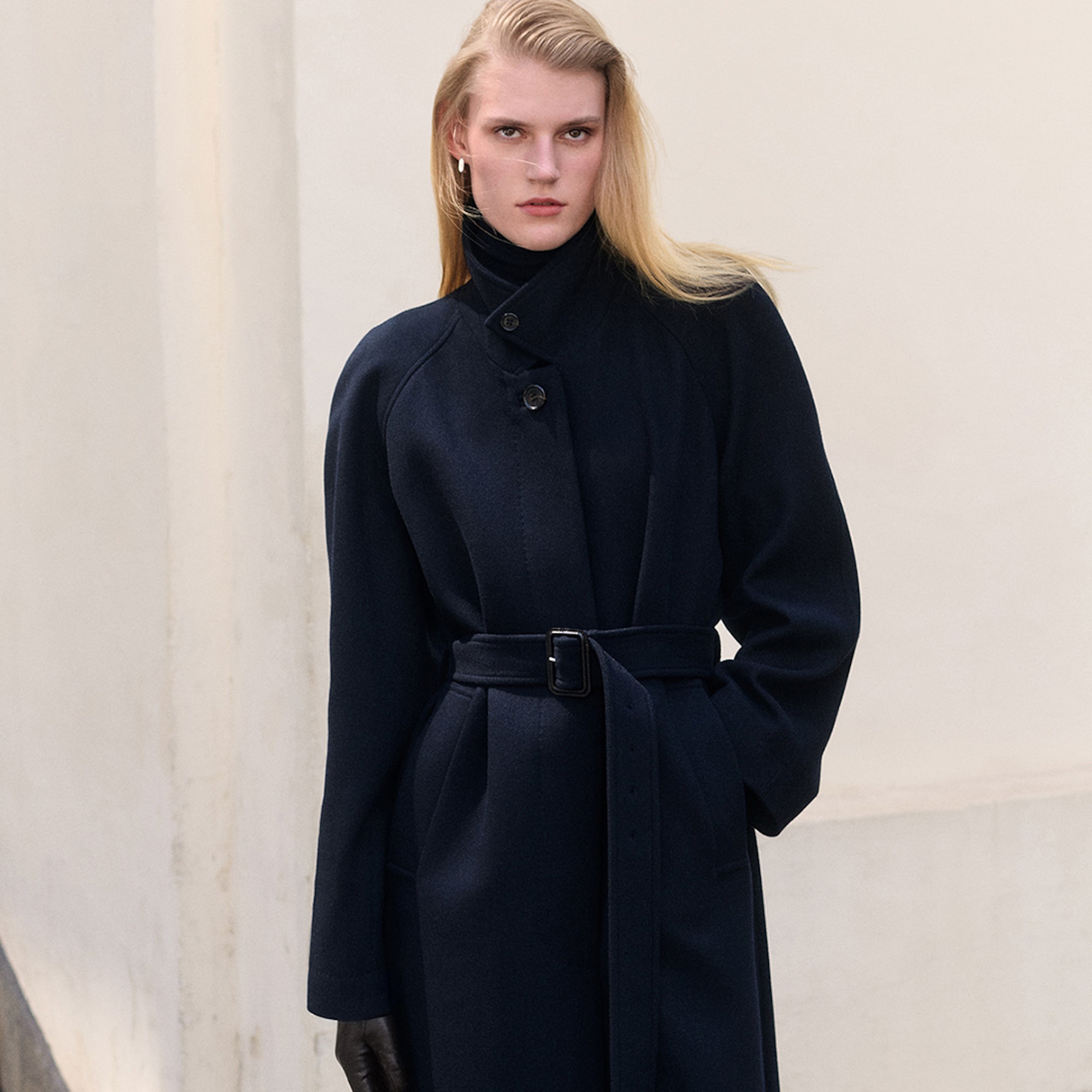 Person wearing a dark, belted trench coat and gloves, standing against a light-colored wall.