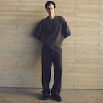 Placeholder for: Person in a cozy, oversized dark sweater and brown trousers stands against a textured beige wall with hands behind their back.