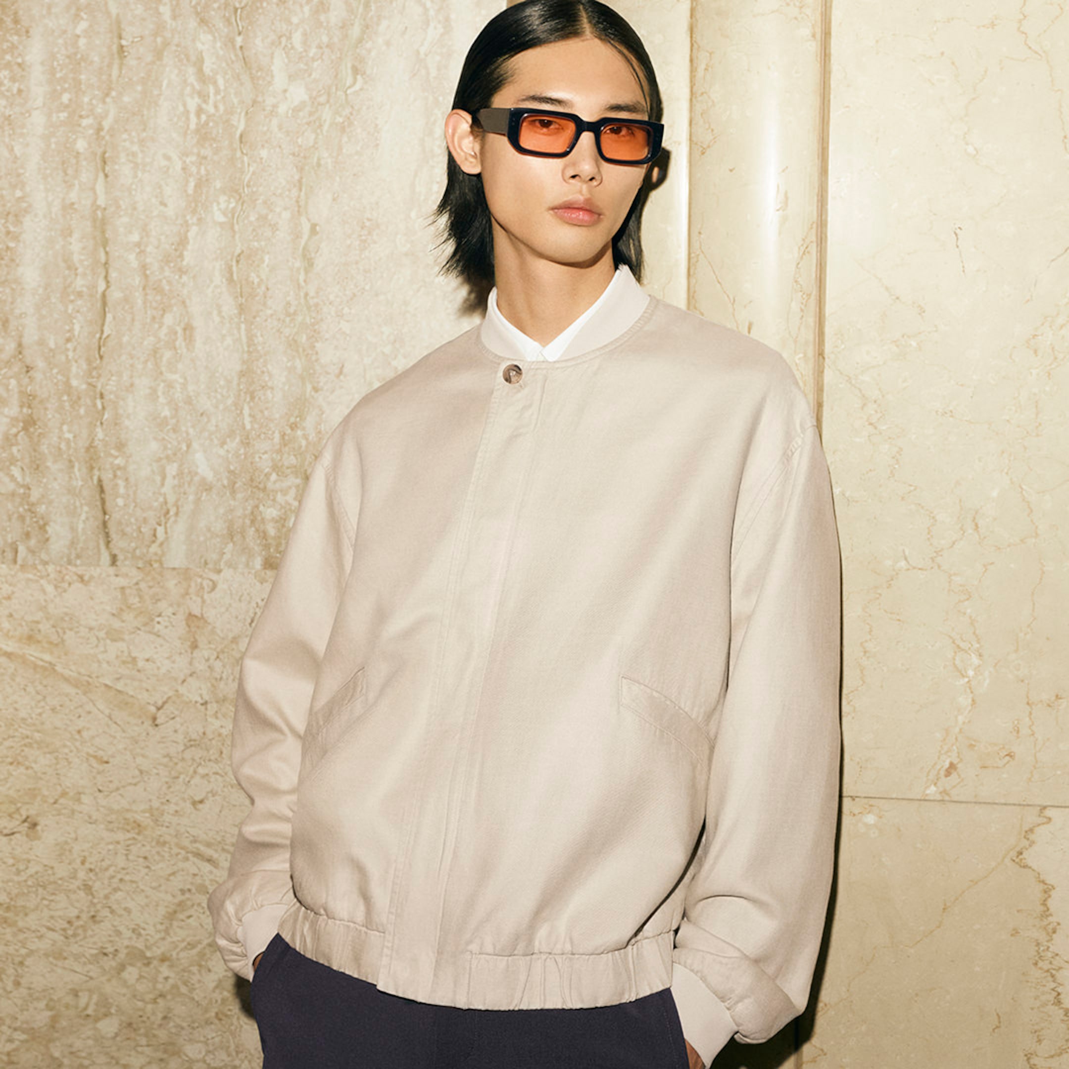 A person with long hair wearing sunglasses and a light beige jacket stands against a marble wall, hands in pockets.