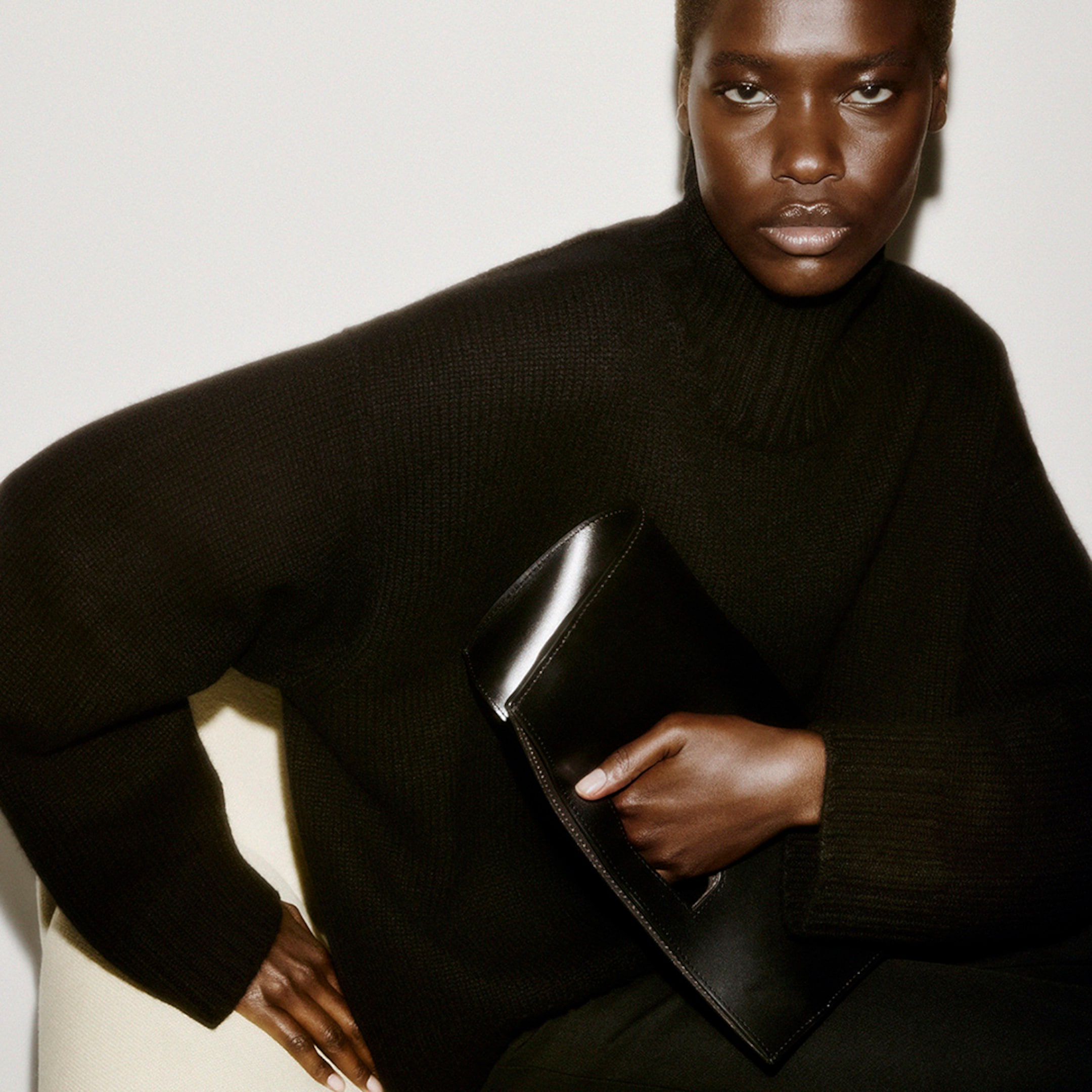 Person in a black turtleneck holds a sleek black clutch. The image has a soft focus and warm lighting.