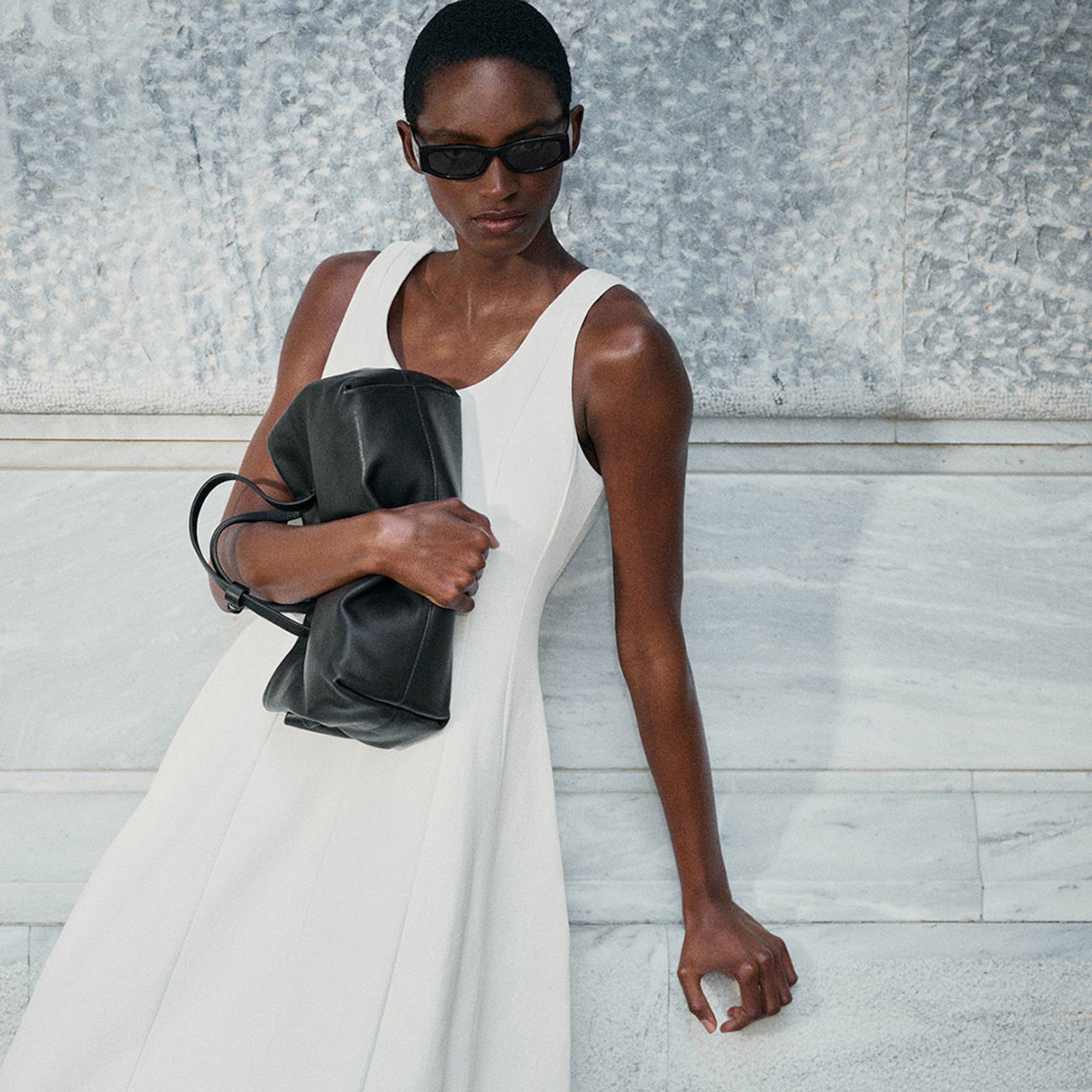 A person in sunglasses wearing a white dress, holding a large black bag, standing against a textured, light-colored wall.