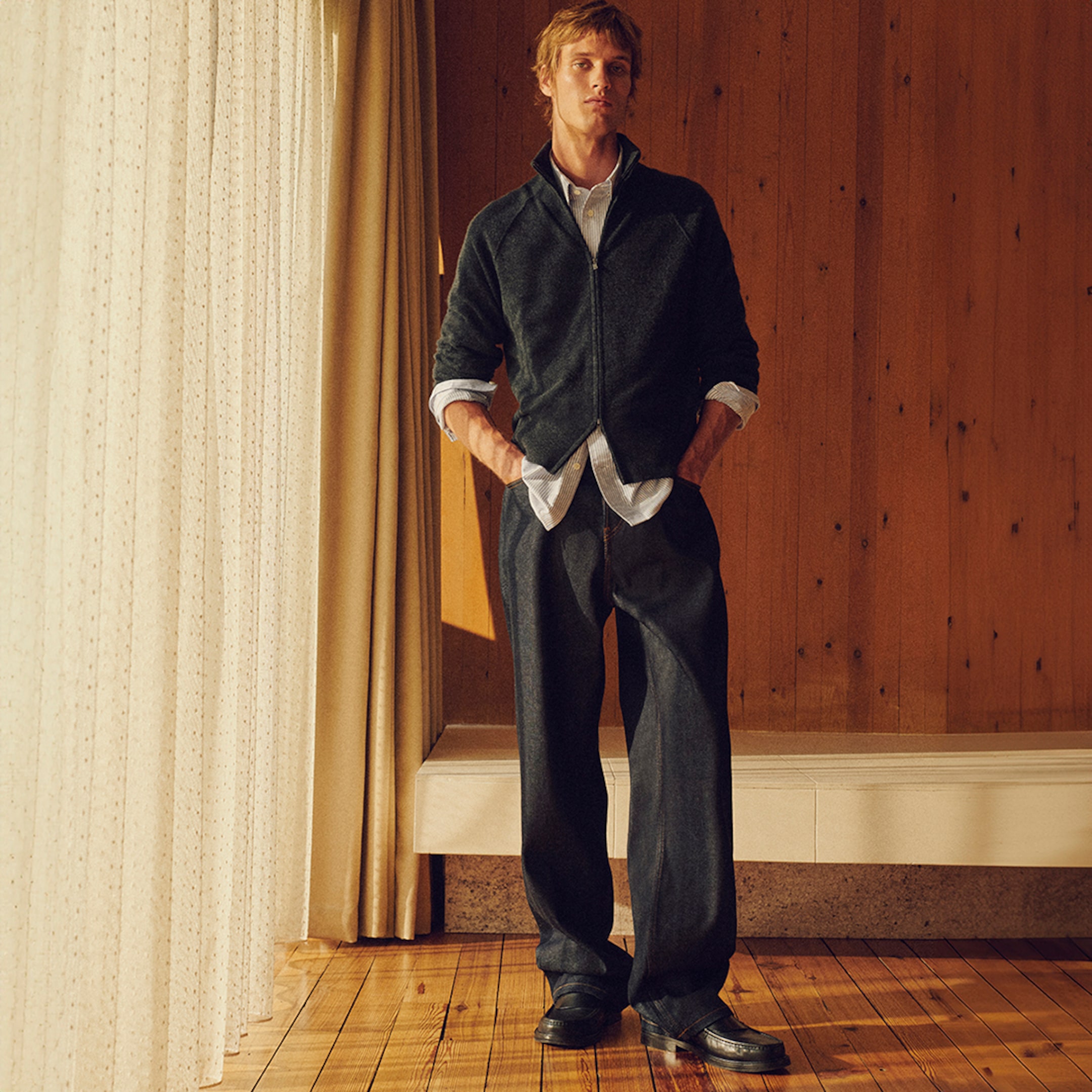 Man in a dark jacket and pants stands in a warmly lit room with wood paneling, hands in pockets, gazing upward.