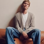 Placeholder for: Person lounging on a brown leather chair, wearing a beige cardigan over a white shirt and blue jeans against a plain wall.