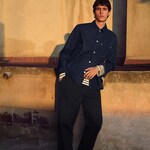 Placeholder for: A person in a denim shirt and black pants leans against a wall in warm sunset light.