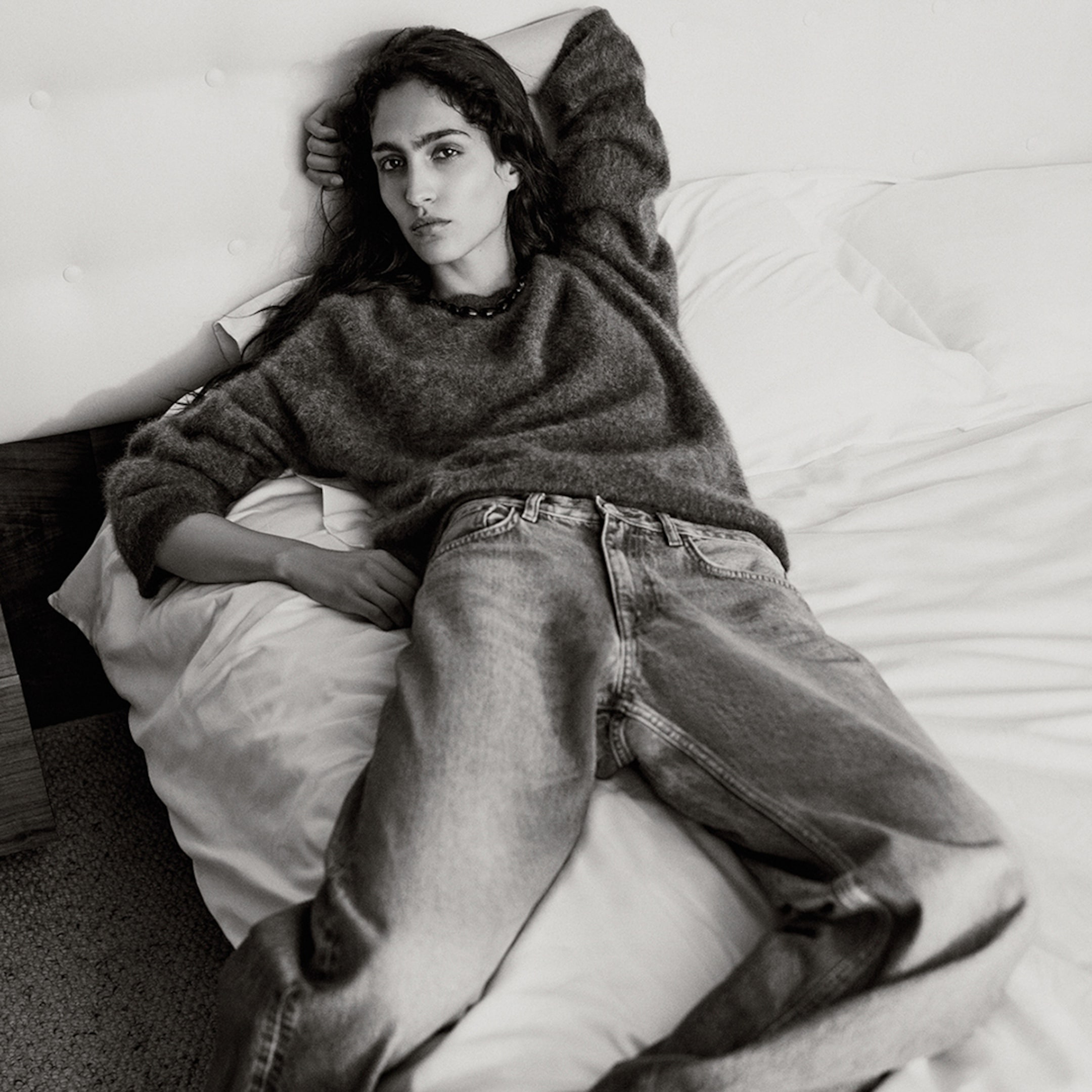Person in a relaxed pose on a bed, wearing a sweater and jeans, with one arm behind their head and a neutral expression. Black and white photo.
