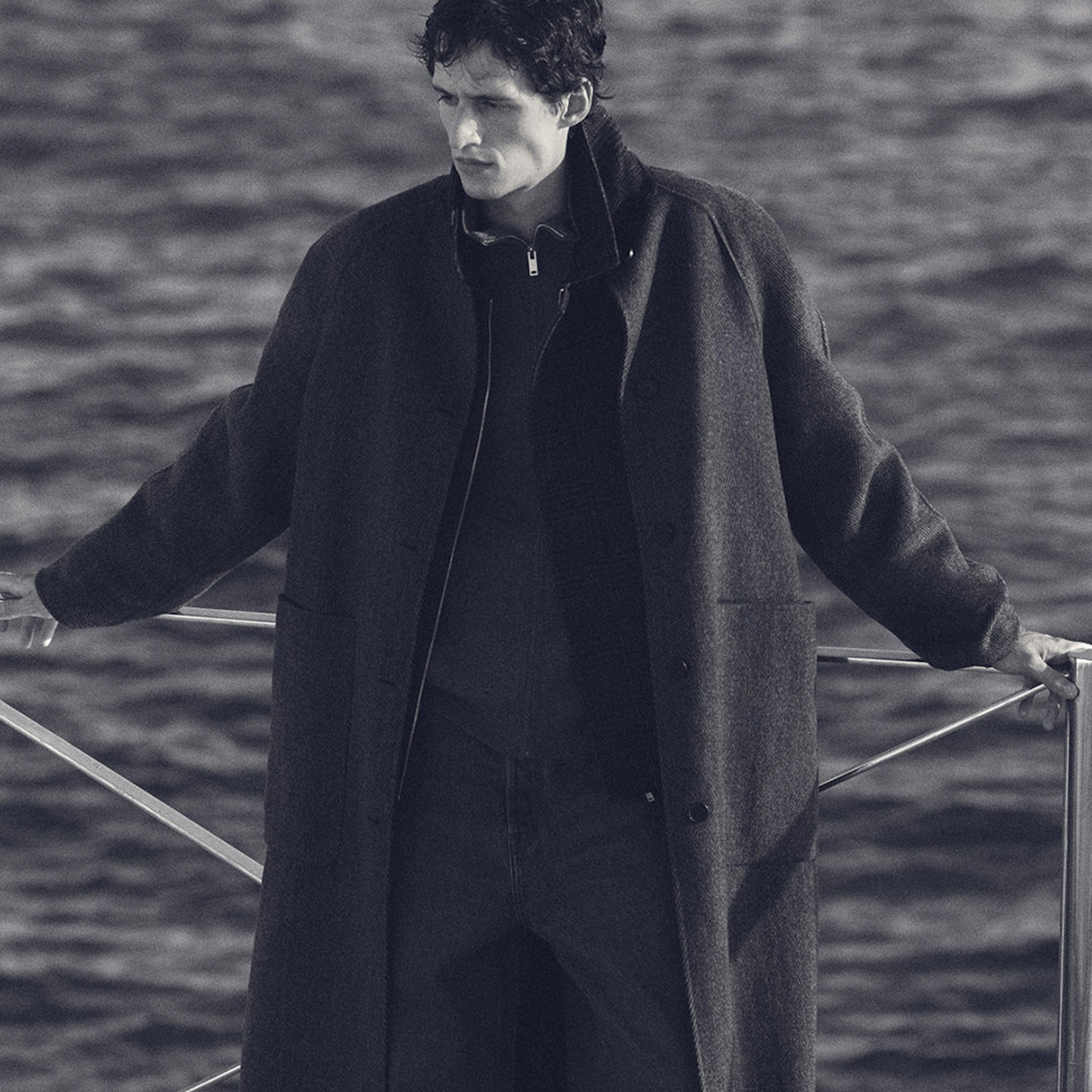 Man in dark overcoat standing by the railing near water, gazing to the side in a monochrome setting.