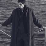 Placeholder for: Man in dark overcoat standing by the railing near water, gazing to the side in a monochrome setting.
