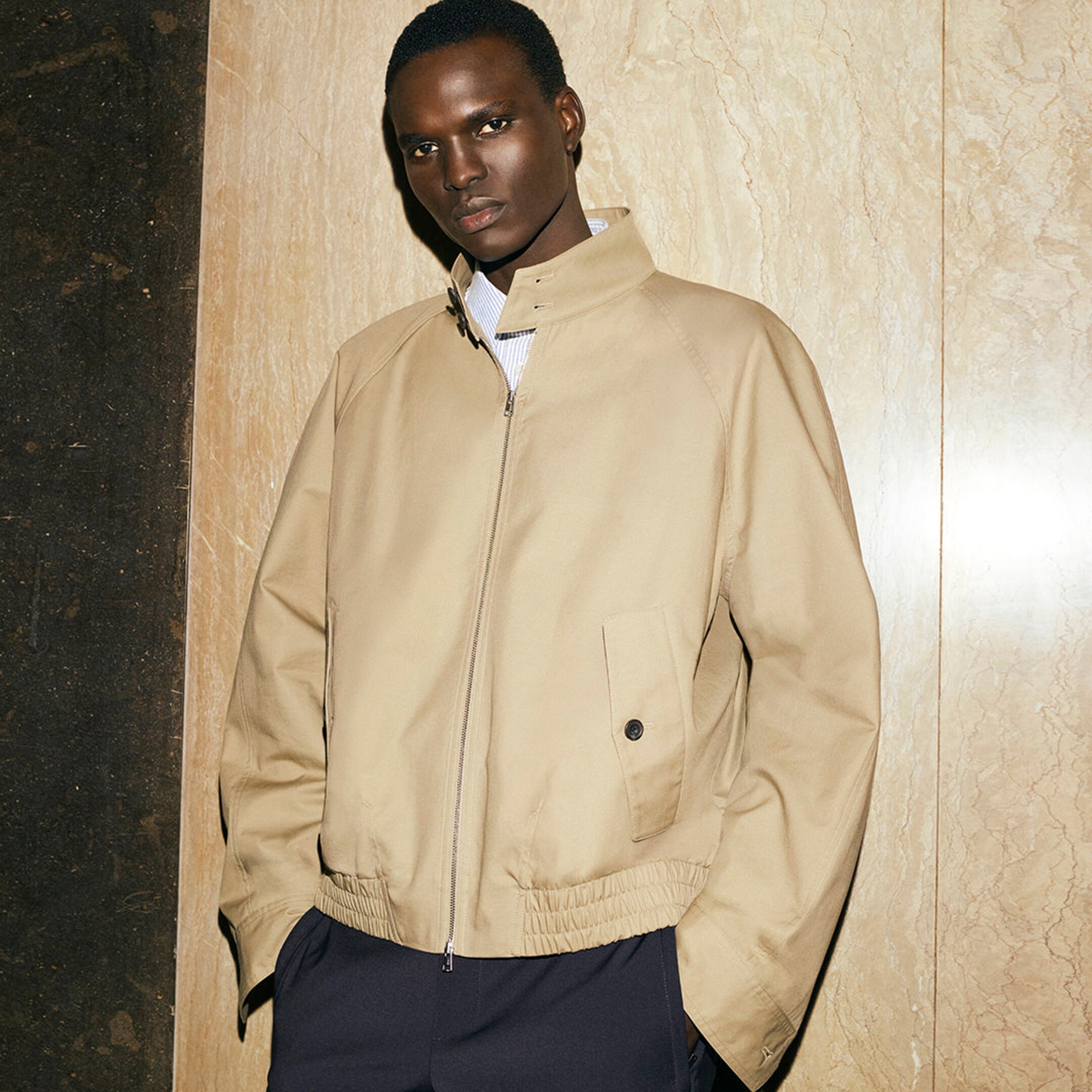 Person wearing a beige jacket over a white shirt, standing against a marble wall with hands in pockets, looking at the camera.