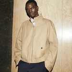 Placeholder for: Person wearing a beige jacket over a white shirt, standing against a marble wall with hands in pockets, looking at the camera.