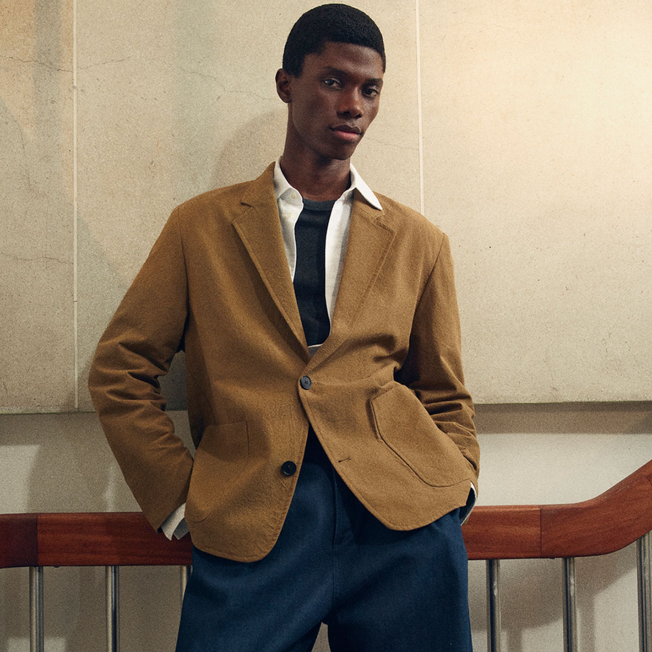 Model wearing a brown blazer over a white shirt and black top, paired with blue trousers, standing against a light-colored wall.