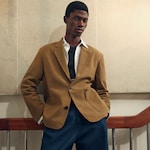 Placeholder for: Model wearing a brown blazer over a white shirt and black top, paired with blue trousers, standing against a light-colored wall.