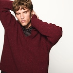 Placeholder for: Person wearing a red knit sweater and striped shirt, posing with arms raised, against a plain white background.