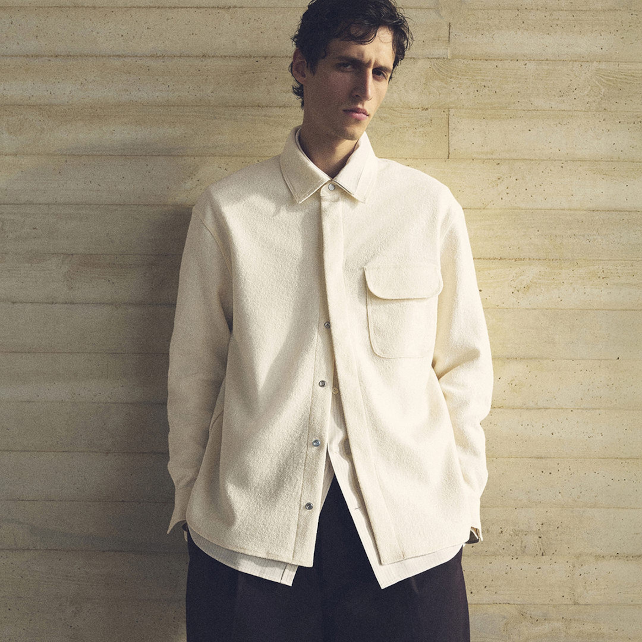 Man wearing a cream textured jacket over a white shirt, standing against a beige textured wall, looking directly at the camera.