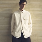 Placeholder for: Man wearing a cream textured jacket over a white shirt, standing against a beige textured wall, looking directly at the camera.