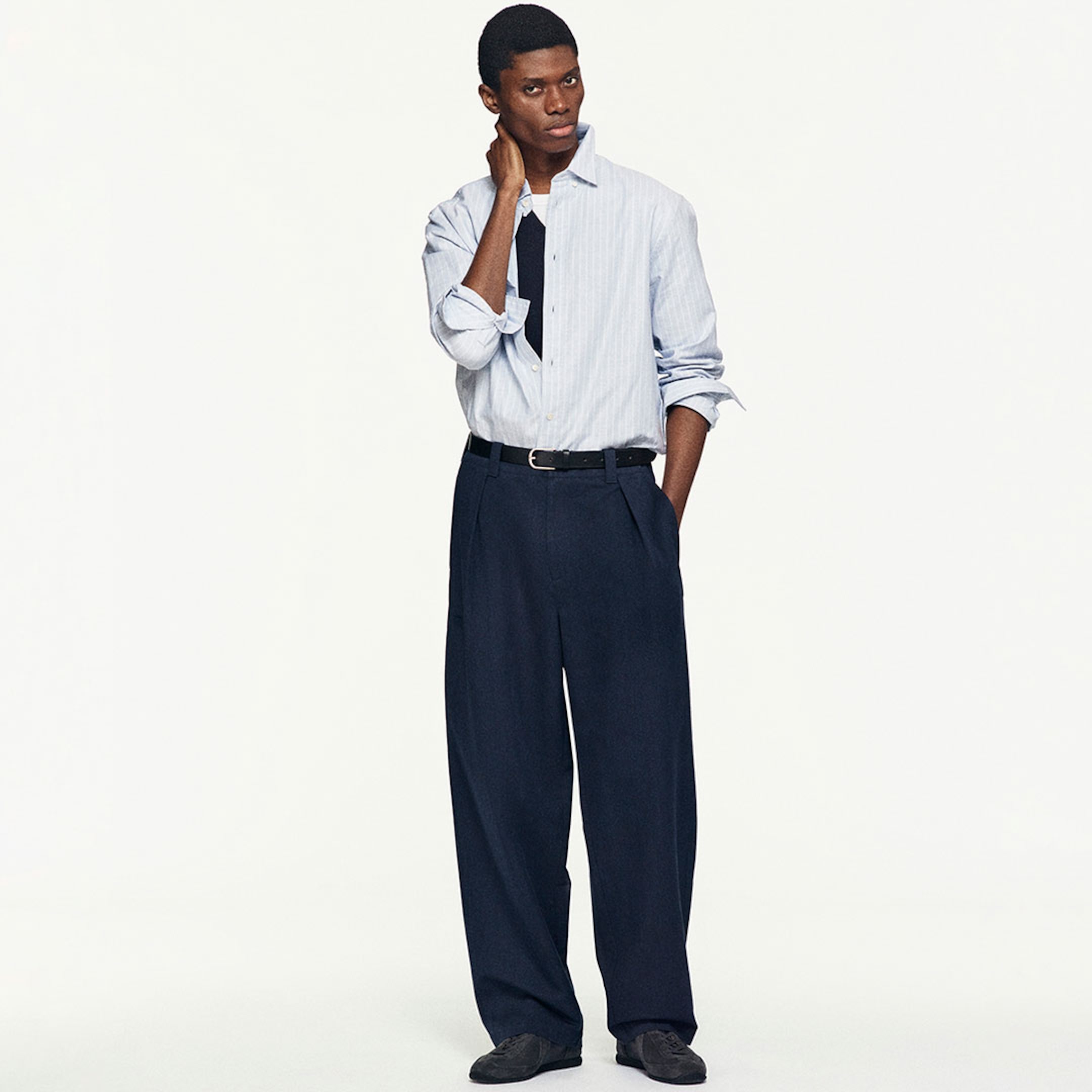 Model wearing a light blue striped shirt over a black top, with navy blue wide-leg trousers and black shoes, standing with one hand on their neck.