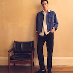 Placeholder for: A man stands by a chair, wearing a blue jacket over a white shirt with black pants and shoes, against a beige wall.