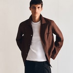 Placeholder for: A person wearing a brown button-up shirt over a white T-shirt and black pants, standing against a neutral background.