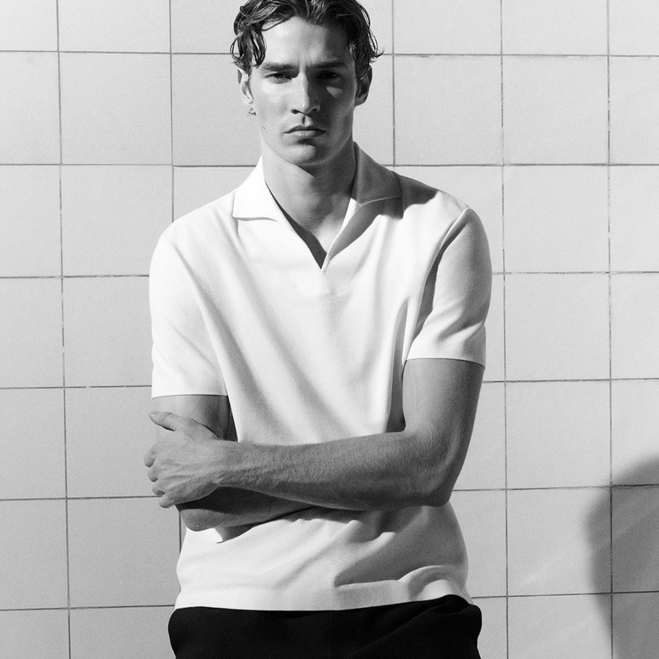 Man in a white short-sleeve shirt stands with arms crossed against a tiled wall, looking ahead with a serious expression. Black and white photo.