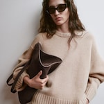 Placeholder for: Woman in a beige sweater holding a suede handbag, wearing large sunglasses, against a plain background.