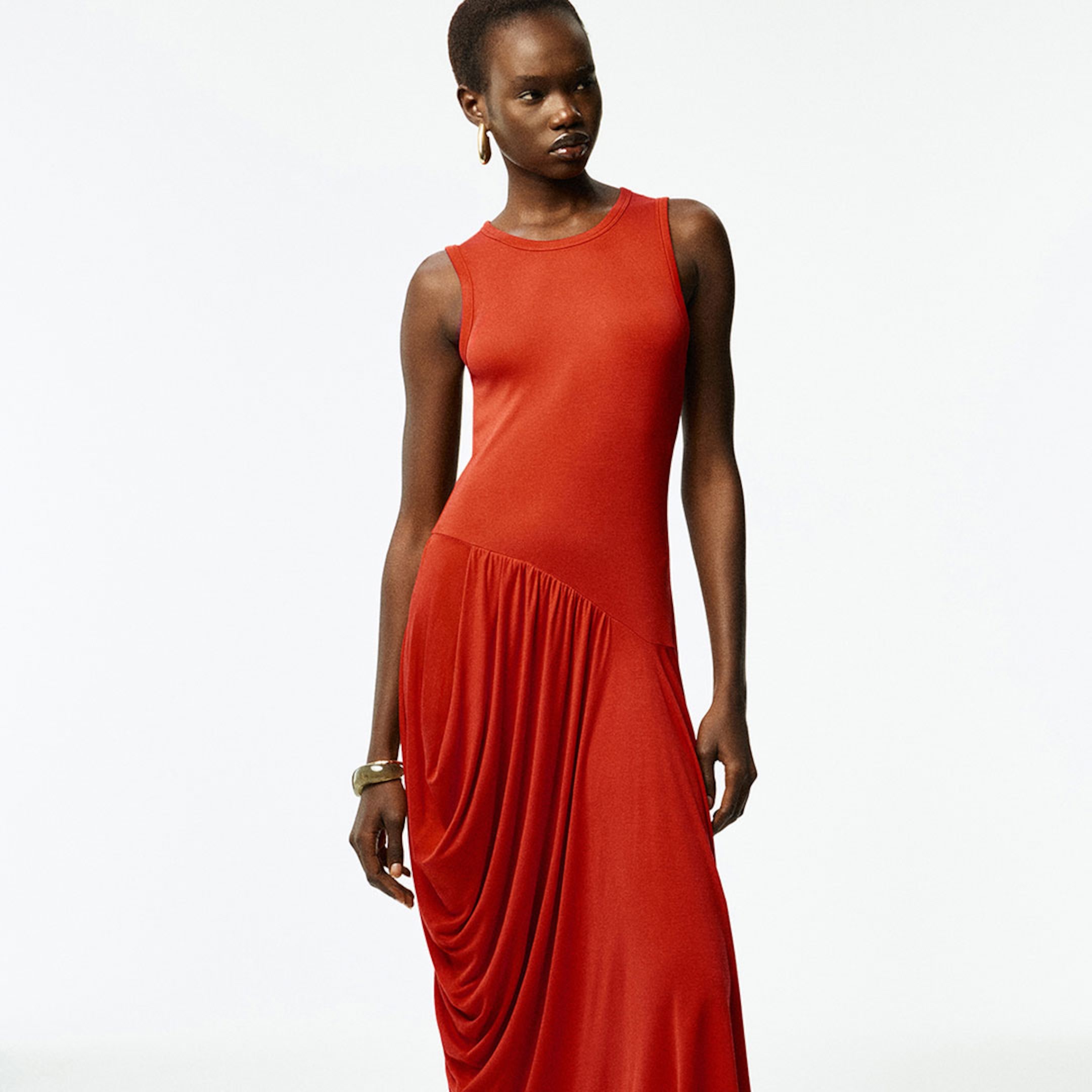 Person wearing a sleeveless, draped, long red dress with gold hoop earrings and a bracelet, standing against a plain white background.