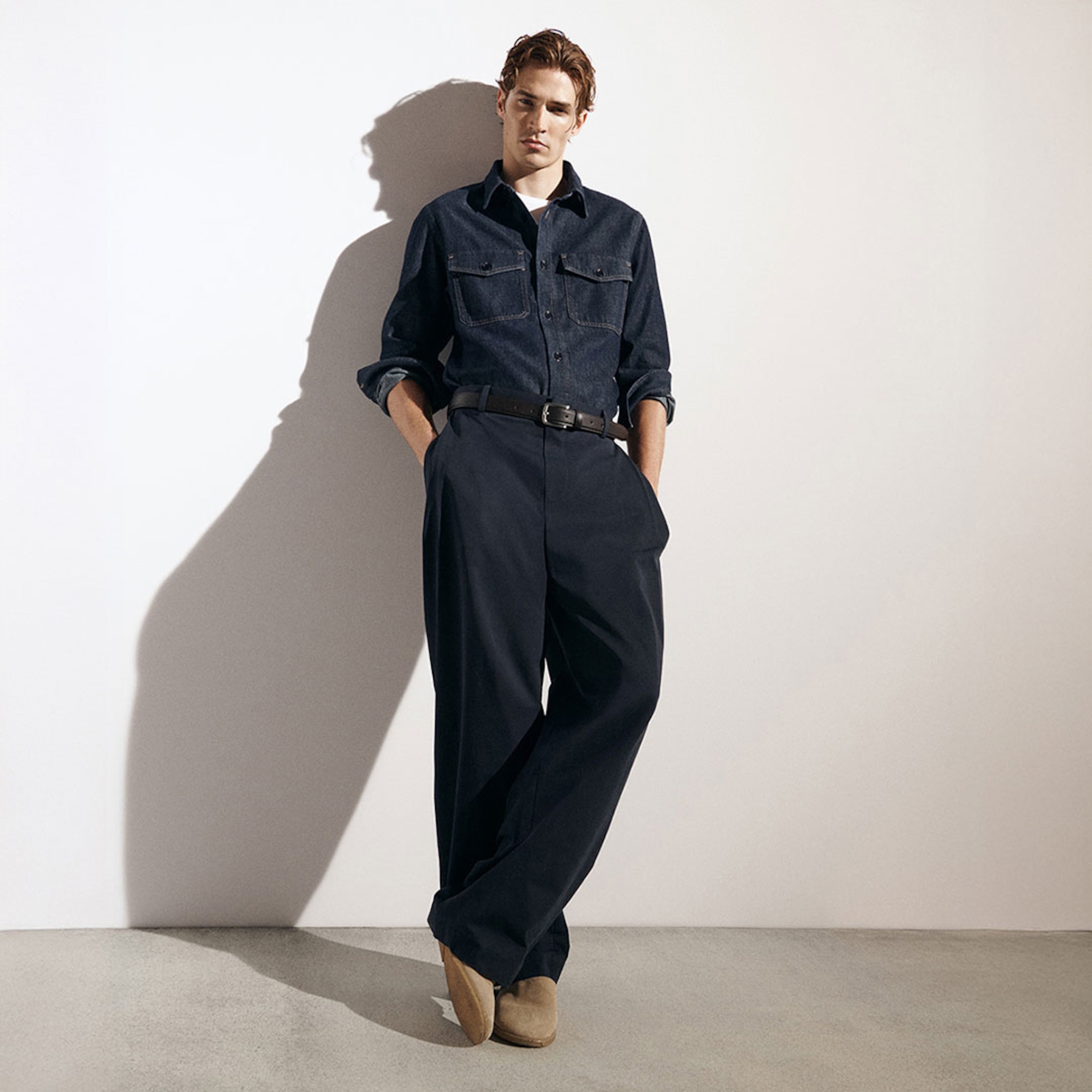 Person wearing a dark denim shirt and wide-leg pants, standing against a white wall with hands in pockets and feet crossed, casting a shadow.