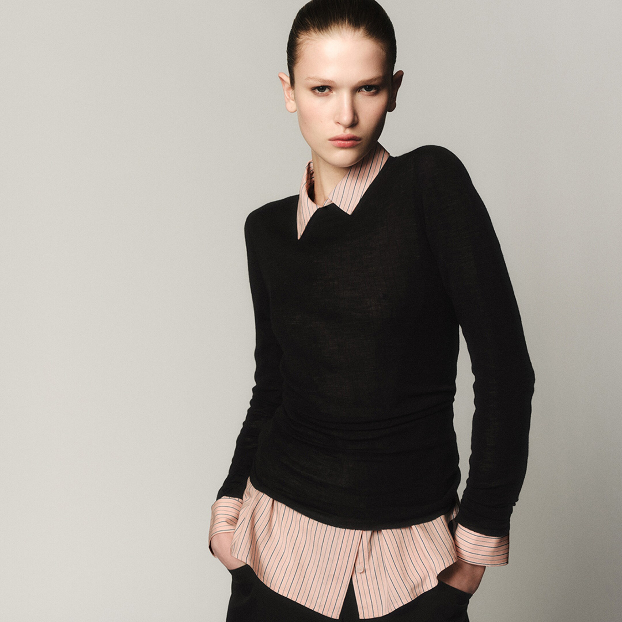 Person wearing a black sweater over a striped shirt, posing with hands in pockets against a neutral background.