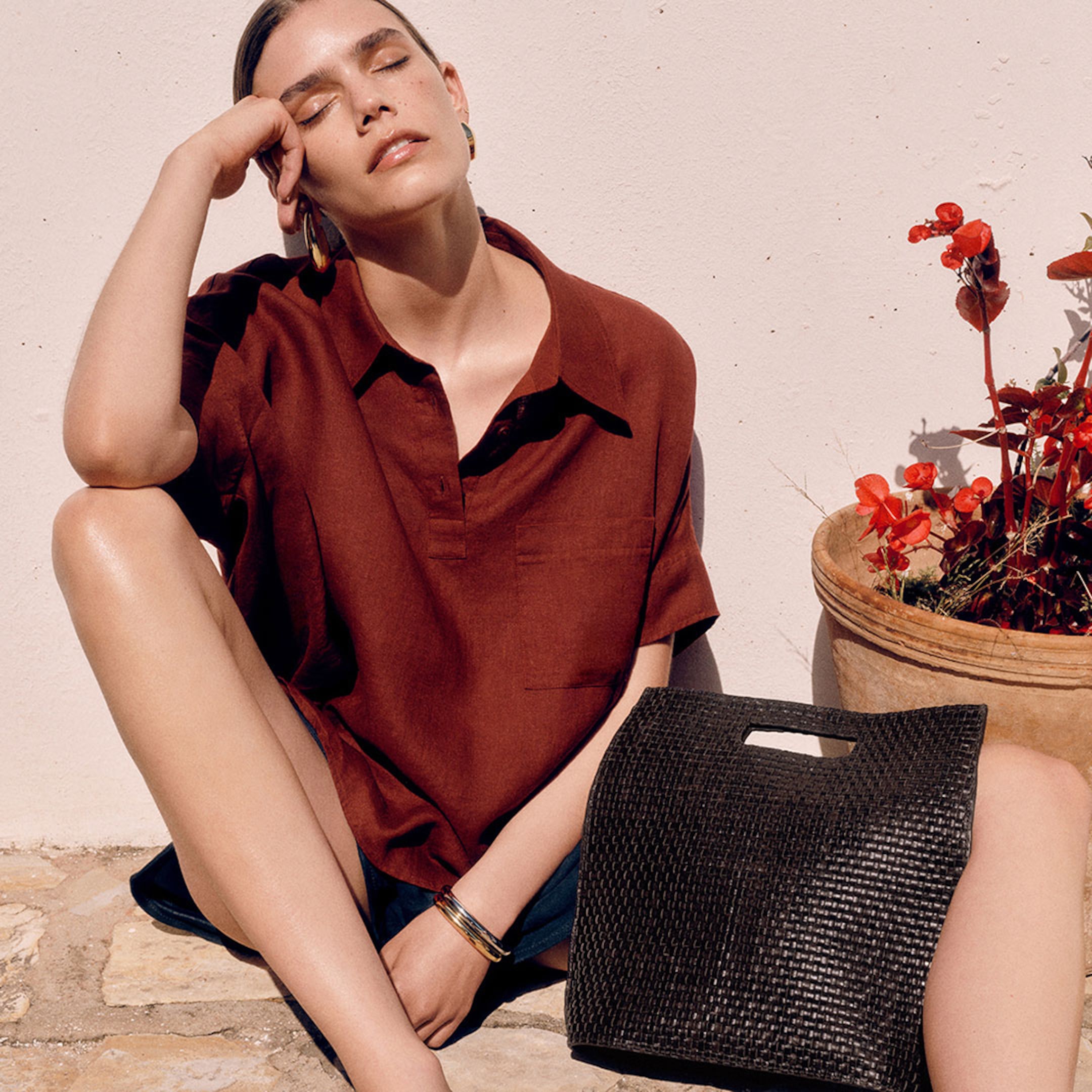 A sunlit profile shot of a woman with long wavy hair sitting outdoors against a white textured wall. She is wearing a bright red, knitted V-neck sleeveless cardigan vest with tonal buttons and black drawstring trousers. The look is accessorized with chunky silver hoop earrings and a thick silver bangle.