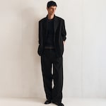Placeholder for: Man wearing a black textured suit, relaxed fit, with matching pants, black shirt, and shiny black shoes, standing against a light background.