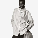 Placeholder for: Person in a stylish, oversized button-up shirt with a unique collar design stands confidently, hand in pocket, against a plain background.