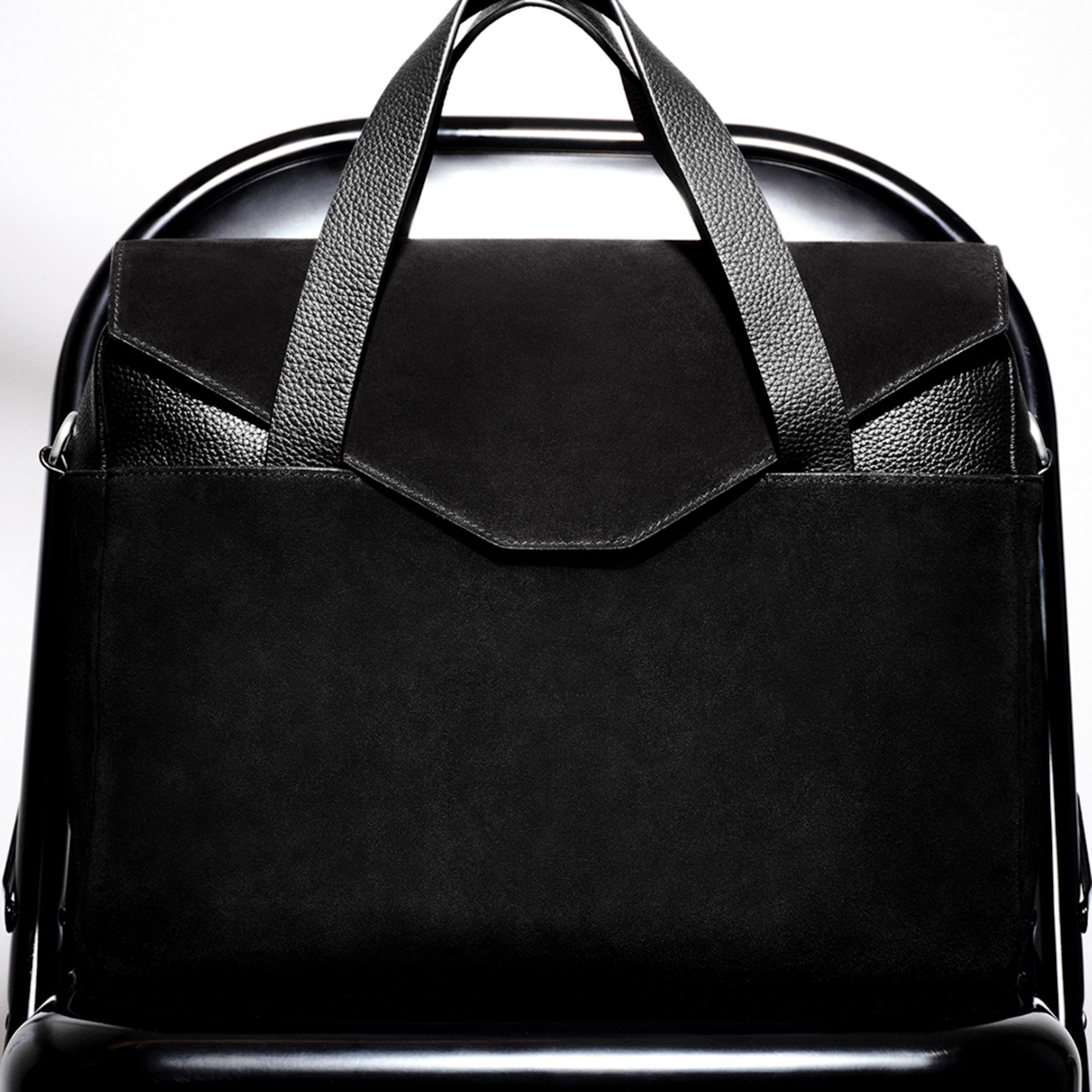 A stylish black leather bag with textured handles rests on a sleek black chair, highlighted against a bright background.