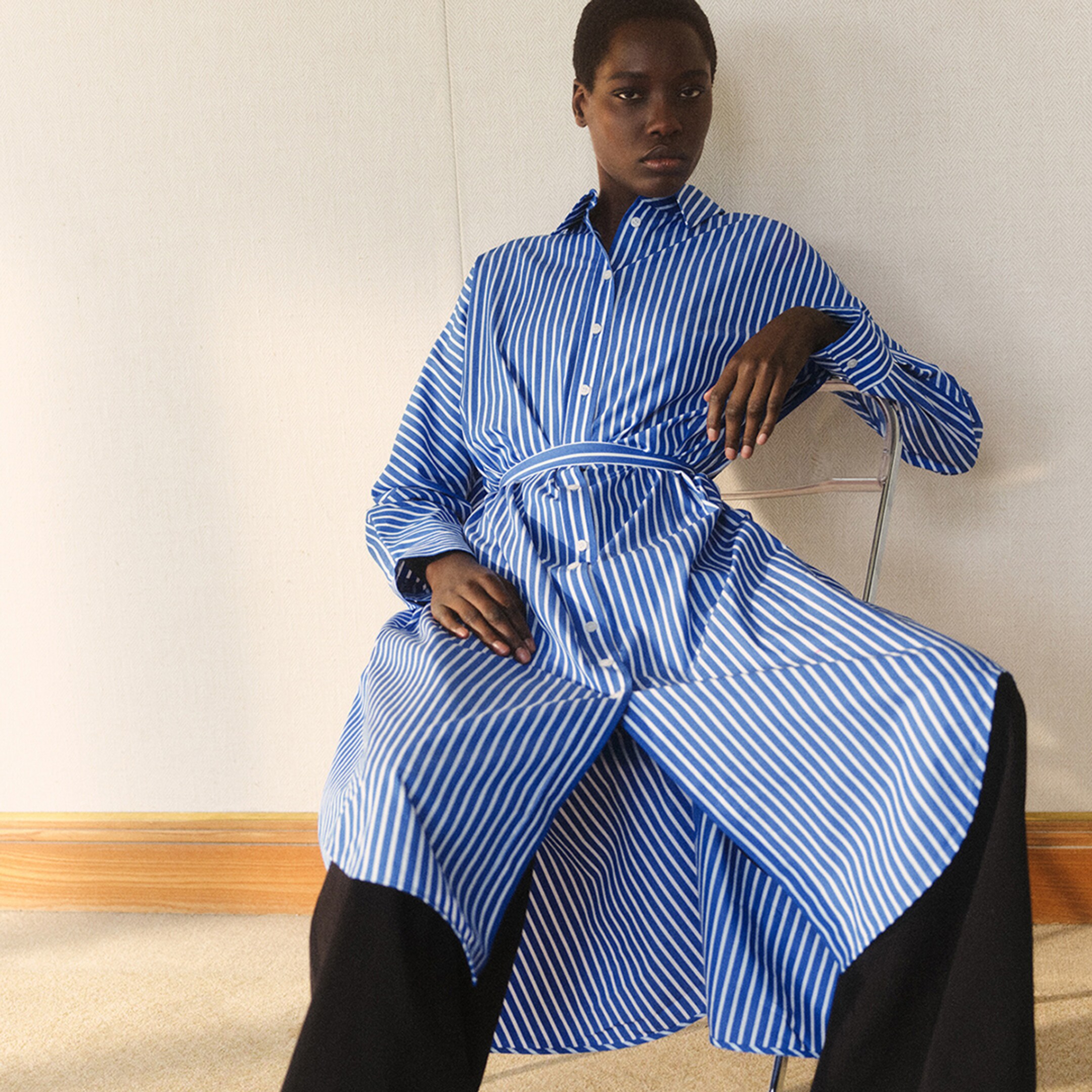 Person sitting on a chair wearing a blue and white striped dress with black pants, against a plain wall and wooden floor.