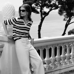 Placeholder for: A black-and-white lifestyle photograph of a woman standing and leaning against a white decorative stone pillar outdoors. She is wearing a striped short-sleeved knit polo shirt and wide-leg white trousers, accessorized with dark cat-eye sunglasses and silver bangles. The background shows a hazy view of the sea.