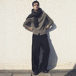 Placeholder for: Person wearing a gray cardigan, black scarf, and wide-leg trousers stands against a mosaic-tiled wall in sunlight.
